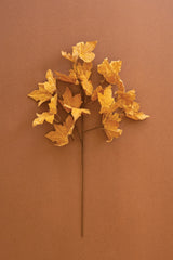 A single stem of artificial maple leaves in warm golden yellow and orange tones, arranged against a solid warm brown background.