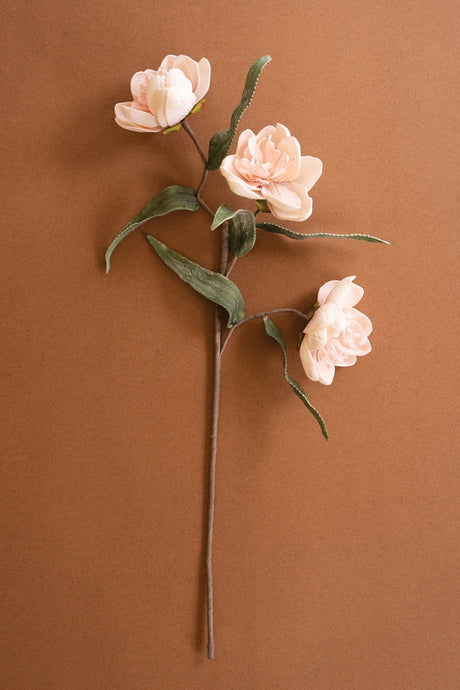 Three pale blush pink artificial flowers with green leaves on a single stem, arranged against a solid warm brown background.