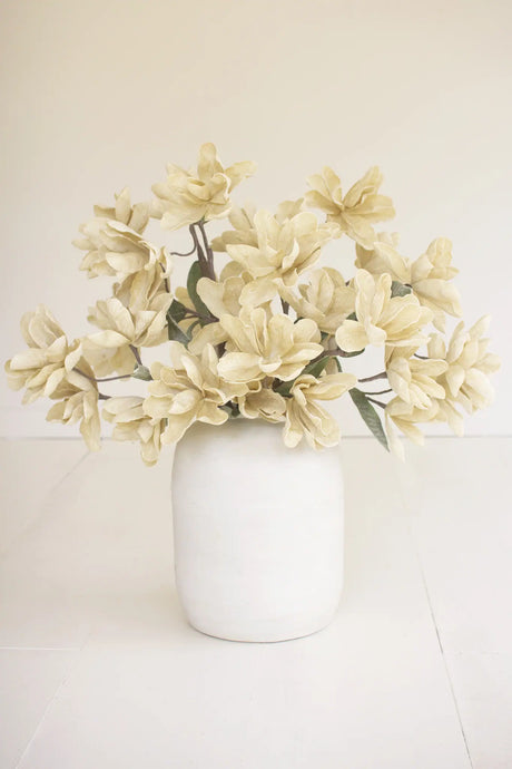 A bouquet of cream-colored artificial magnolia flowers arranged in a matte white ceramic vase, set against a clean, off-white background.