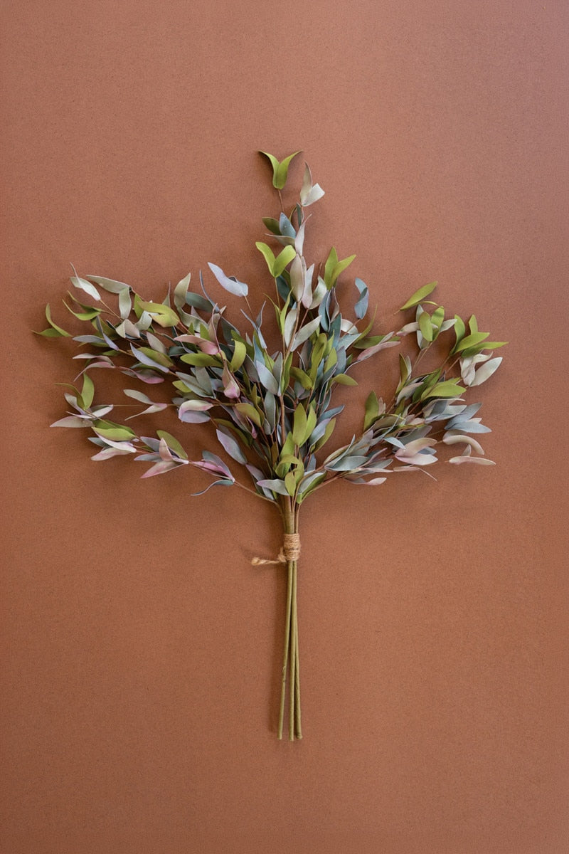 A dried artificial eucalyptus branch bundle with muted green, gray, and soft pink leaves tied with natural twine, displayed against a solid terracotta brown background.