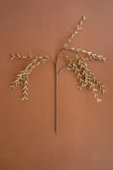 A dried decorative branch with delicate pale beige leaves on a long brown stem, arranged symmetrically against a solid warm terracotta background.