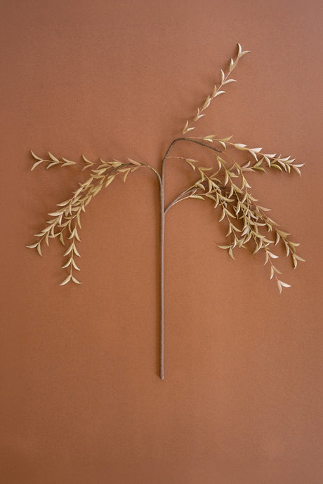 A dried decorative branch with delicate pale beige leaves on a long brown stem, arranged symmetrically against a solid warm terracotta background.