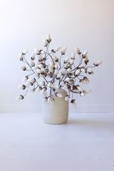 A decorative artificial branch arrangement with white flower buds on a dark brown stem, displayed in a matte off-white ceramic vase against a clean white background.