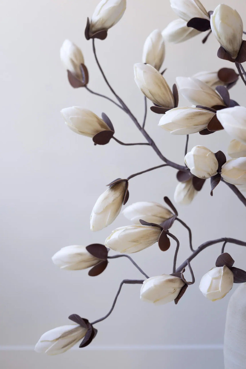 Artificial magnolia flower branches with cream-colored blooms and dark brown leaves on a light neutral background.