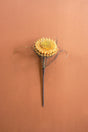 A single dried artificial flower stem with a round, textured golden-yellow bloom and thin dark wire-like accents, centered on a warm terracotta background.