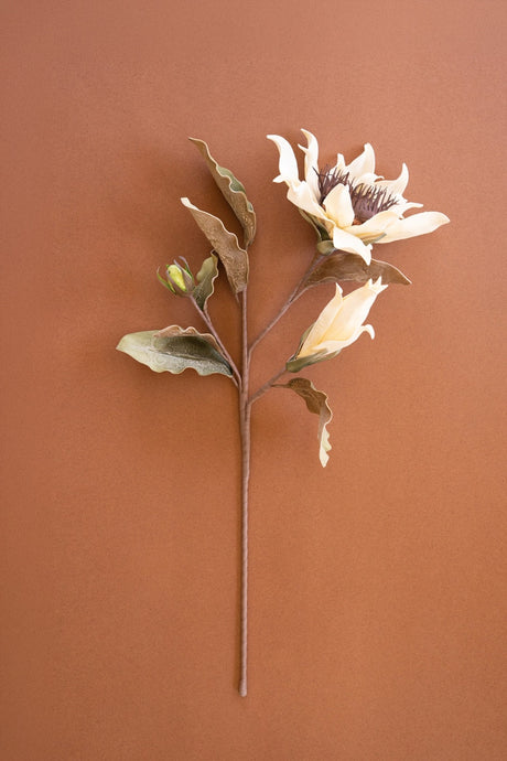 A single artificial flower stem with cream-colored petals, green and brown leaves, and a small bud, displayed against a solid terracotta background.