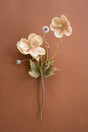 A single artificial flower stem with three pale peach blossoms, green stems and leaves, and small buds, arranged on a warm brown background.