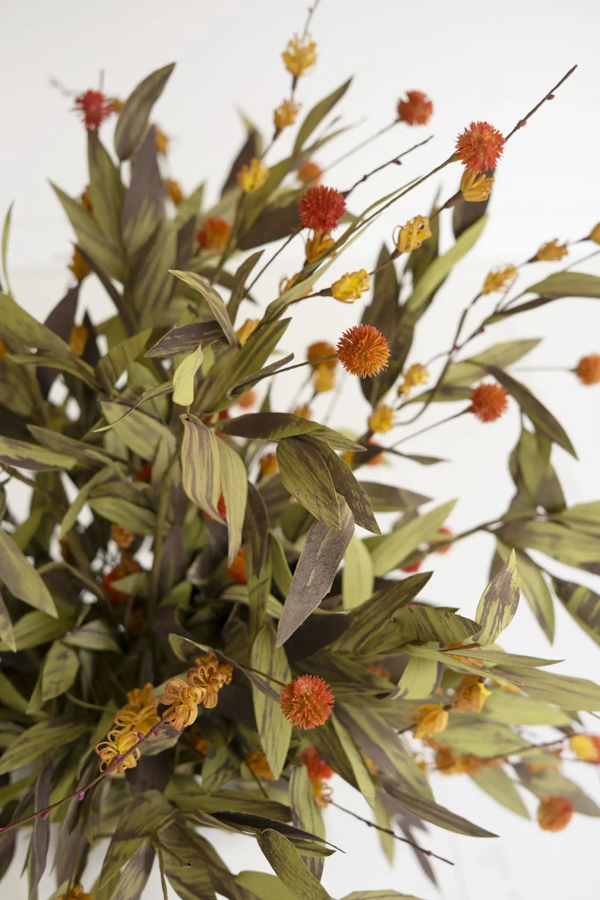 Artificial foliage arrangement with olive green leaves, muted brown stems, and small orange and yellow pom-pom flowers against a plain white background.