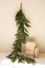 A long green artificial evergreen garland hangs from a brown string against a plain cream wall, with a small stack of old books on a white surface below.