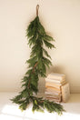 A long green artificial evergreen garland hangs from a brown string against a plain cream wall, with a small stack of old books on a white surface below.