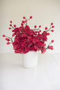 A red artificial floral arrangement with clusters of ruffled blossoms and small round berries, displayed in a white cylindrical vase against a clean white background.