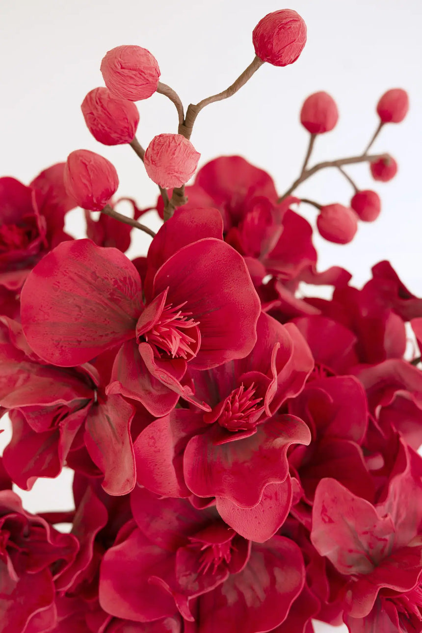 A close-up of a bouquet of artificial red orchid flowers with multiple pinkish-red buds on thin brown stems, set against a plain light background.