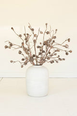 A white ceramic vase holding a dried botanical arrangement with brown branches and small seed pods, set against a plain off-white wall and floor.