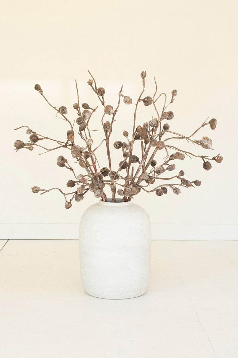 A white ceramic vase holding a dried botanical arrangement with brown branches and small seed pods, set against a plain off-white wall and floor.