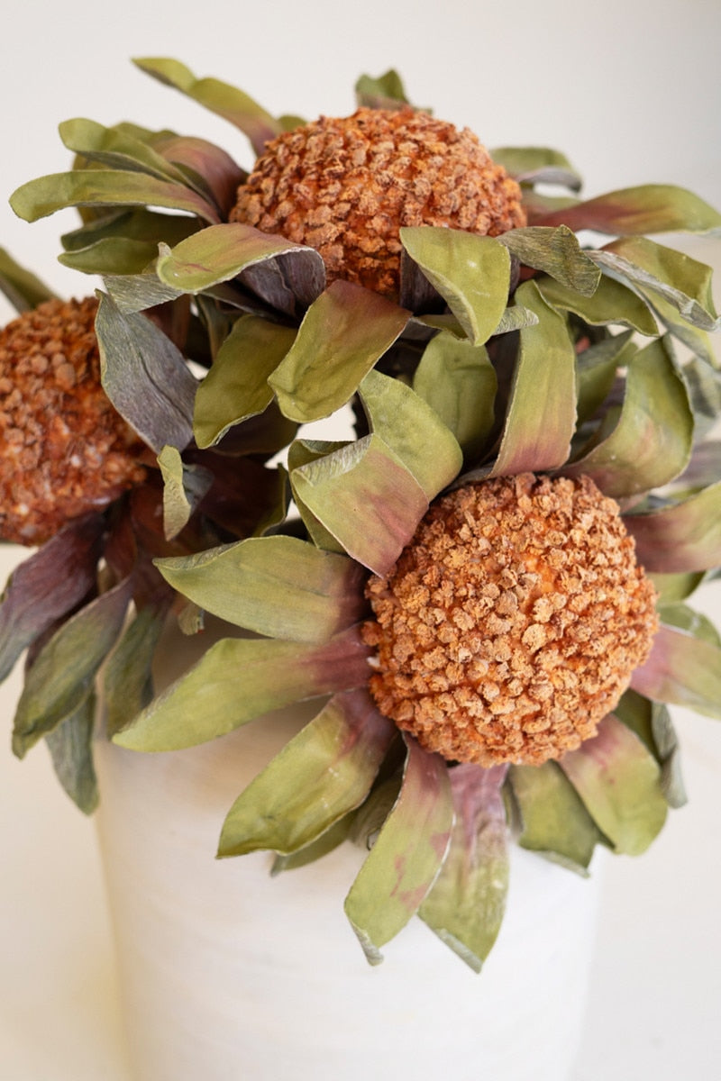 A close-up of a decorative artificial floral arrangement featuring textured orange-brown globe blooms surrounded by olive green and muted burgundy leaves, set in a white container against a plain light background.