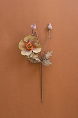 A single dried flower stem with one open pale yellow-brown flower, an orange center, two small buds, and several leaves, arranged against a solid warm brown background.