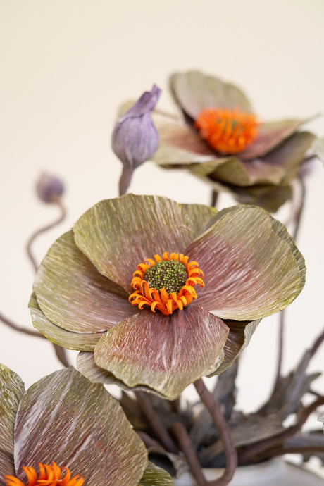 A close-up of artificial paper flowers with textured olive-green and dusty rose petals, bright orange centers, and purple buds on a soft beige background.
