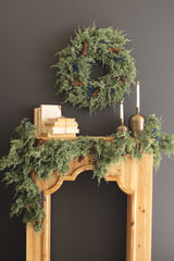 Faux evergreen wreath with dark blue berries and pine cones hanging above a mantle