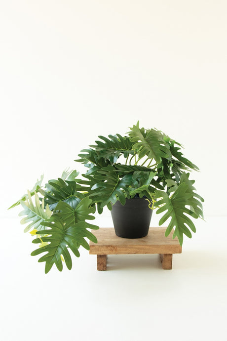 faux split palm plant featuring broad tropical-inspired leaves and a simple black pot standing on small wood pedestal