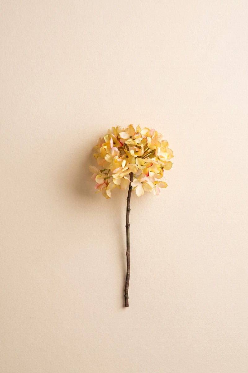 A single artificial hydrangea stem with a round cluster of pale yellow and light pink blossoms on a dark brown stem, photographed against a plain warm beige background.