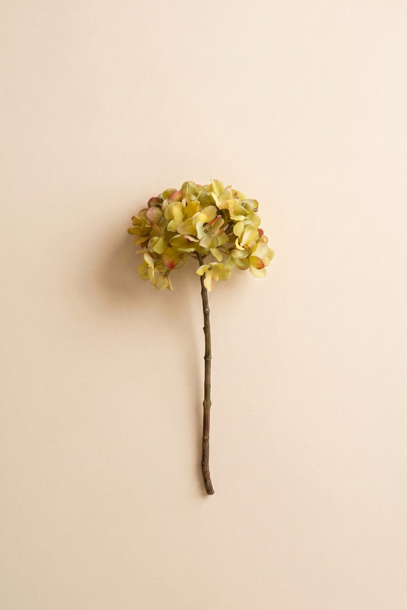 A single artificial flower stem with a round cluster of pale yellow-green hydrangea-like blooms on a thin brown stem, centered against a plain warm beige background.