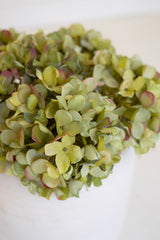 A close-up of a dense cluster of artificial hydrangea-style flowers in soft yellow-green tones with muted pinkish-red edges, arranged in a white container against a clean white background.