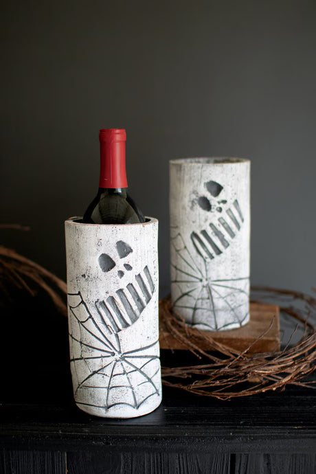Wine bottle in a decorative clay holder with skull face and spider web design against a dark background.