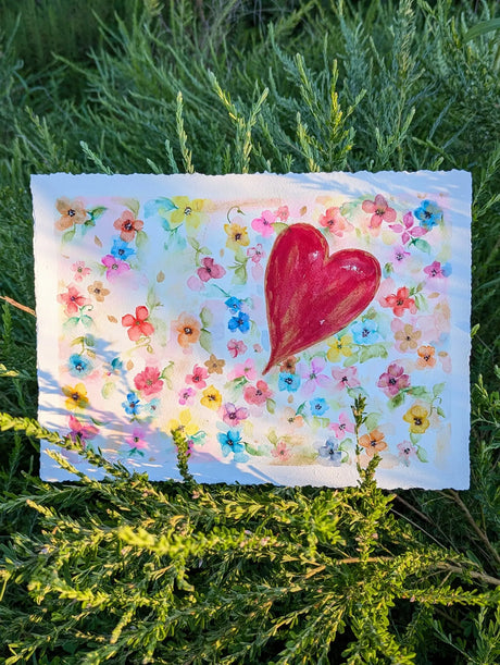 Original watercolor painting featuring a large red heart surrounded by small colorful flowers on a white background placed outdoors on green foliage.
