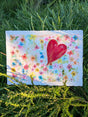 Original watercolor painting featuring a large red heart surrounded by small colorful flowers on a white background placed outdoors on green foliage.
