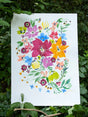 An original watercolor painting featuring a vibrant watercolor floral design with pink, blue, yellow, orange and purple blossoms, displayed outdoors against a background of green leaves and foliage.