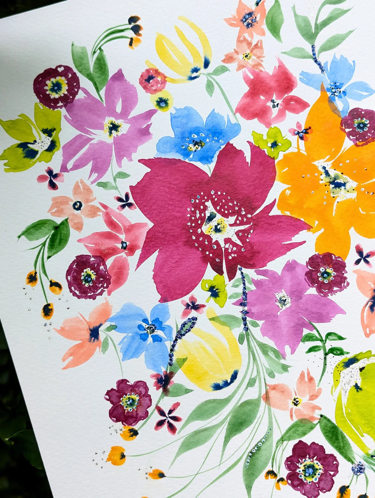 A close-up of a watercolor floral painting on white paper, featuring large and small blossoms in pink, magenta, blue, yellow, orange, purple, and green with delicate leaves and stems.