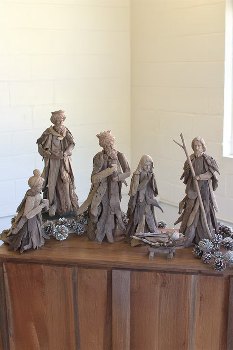 A rustic nativity scene carved from weathered driftwood, displayed on a wooden cabinet against a light-colored brick wall.