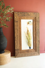 Decorative wooden frame with mirror on a wall next to a black vase and plant.