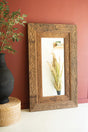 Decorative wooden frame with mirror on a wall next to a black vase and plant.