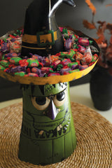 Halloween-themed candy holder with candies and a witch hat on a woven mat.