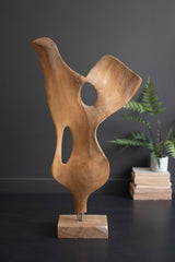 Wooden abstract sculpture on a dark surface with a plant in the background