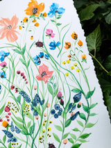 A close-up of a white sheet of paper featuring a colorful watercolor illustration of wildflowers, green leaves, and small insects, with dark green foliage visible along the right edge.