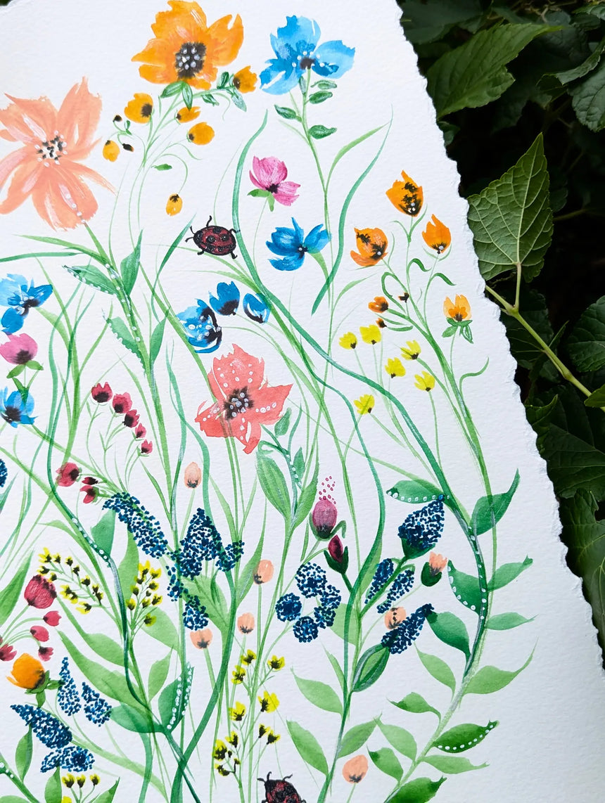 A close-up of a white sheet of paper featuring a colorful watercolor illustration of wildflowers, green leaves, and small insects, with dark green foliage visible along the right edge.