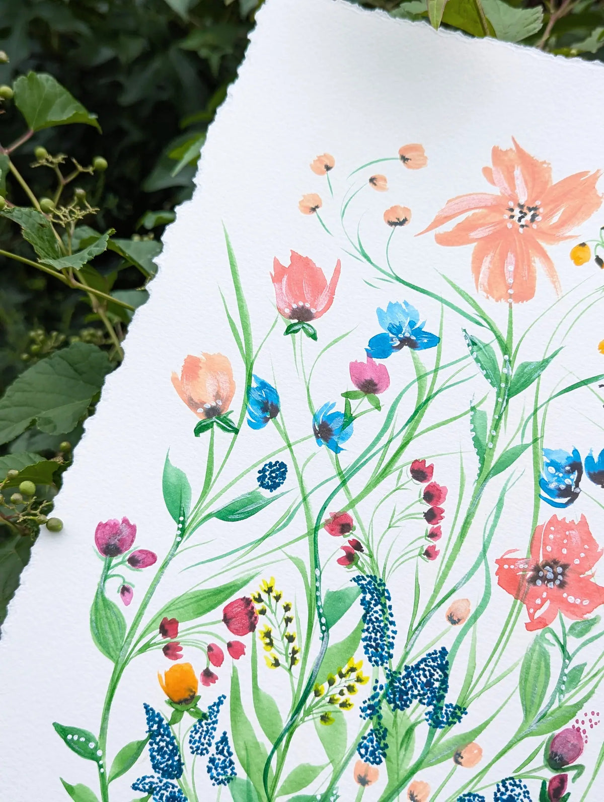 A close-up of a white sheet of paper with hand-painted wildflowers, photographed outdoors against a background of green leaves and vines.