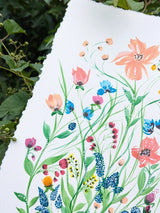 A close-up of a white sheet of paper with hand-painted wildflowers, photographed outdoors against a background of green leaves and vines.