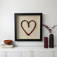 A framed heart-shaped textile wall art piece with a black frame and cream backing, displayed on a white shelf against a white wall, styled with books, a bowl, and two vases.