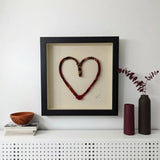 A framed heart-shaped textile wall art piece with a black frame and cream backing, displayed on a white shelf against a white wall, styled with books, a bowl, and two vases.