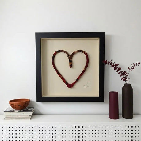 A framed heart-shaped textile wall art piece with a black frame and cream backing, displayed on a white shelf against a white wall, styled with books, a bowl, and two vases.