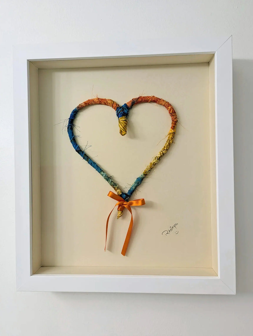 multi-colored recycled fabric heart in white wood frame behind museum glass