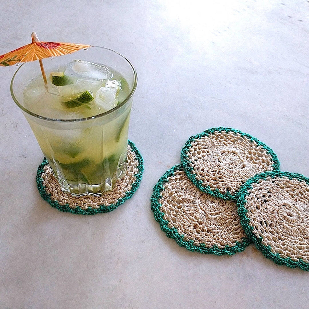 set of four round crochet cotton yarn coasters in natural tan color and teal border - one shown with umbrella drink on top