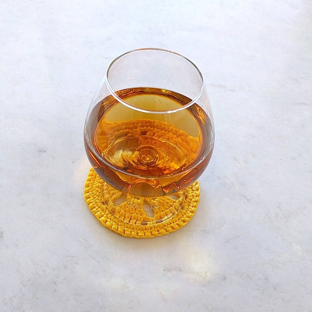 single bright yellow crocheted coaster shown with cognac filled glass